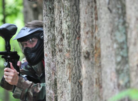 a person with a paintball gun peeking out from the back of a tree