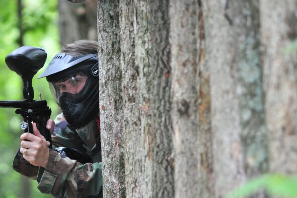 a person with a paintball gun peeking out from the back of a tree