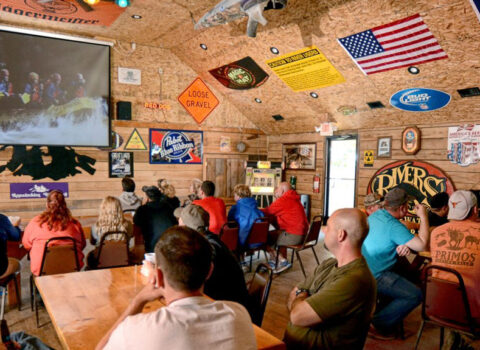 a group of people in the bar looking at photos from a rafting trip on the screen