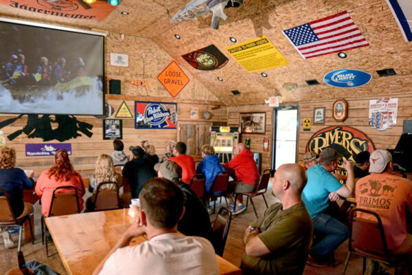 a group of people in the bar looking at photos from a rafting trip on the screen